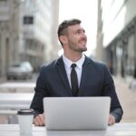 Smiling businessman in formal attire working on a laptop outdoors, exuding confidence and happiness.